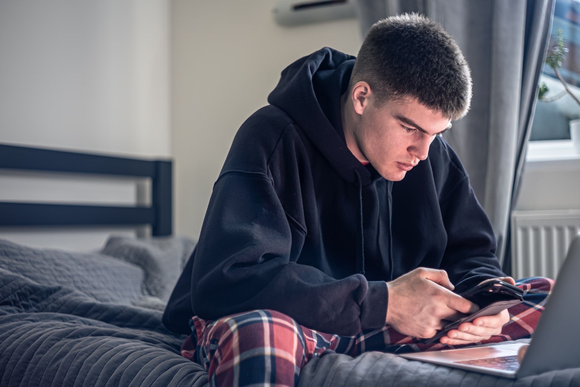 A teenager guy sits in a room on a bed and uses a laptop.